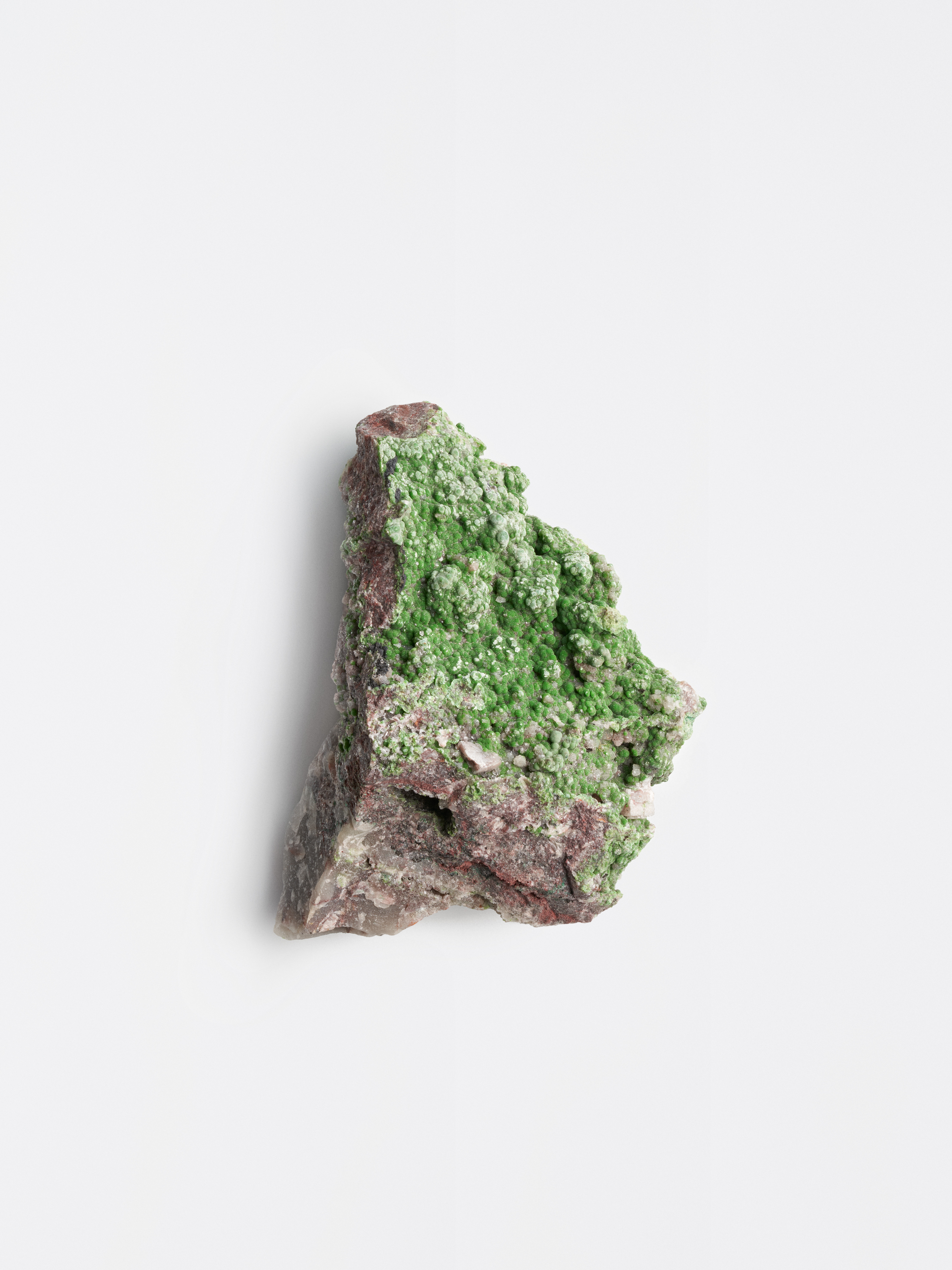 Grünes Mineral auf braun-grauem Malachit-Gestein vor hellem Hintergrund.