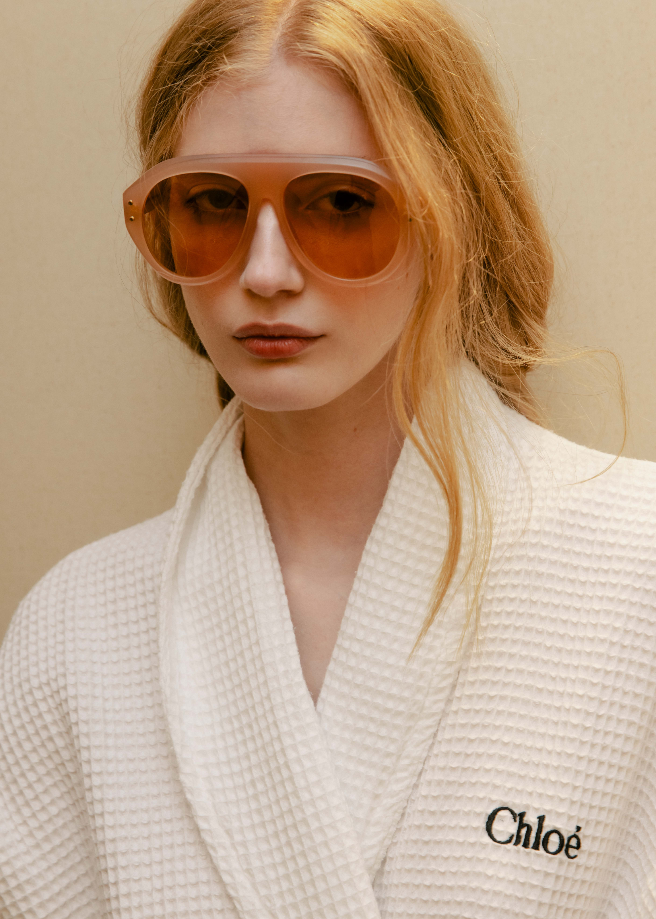 Frau mit rötlichen Haaren trägt eine auffällige orangene Sonnenbrille und einen weißen Bademantel mit der Bestickung "Chloé".
